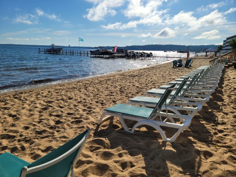 A beach scene with lounge chairs on the sand, a pier in the water, and a blue sky with clouds.