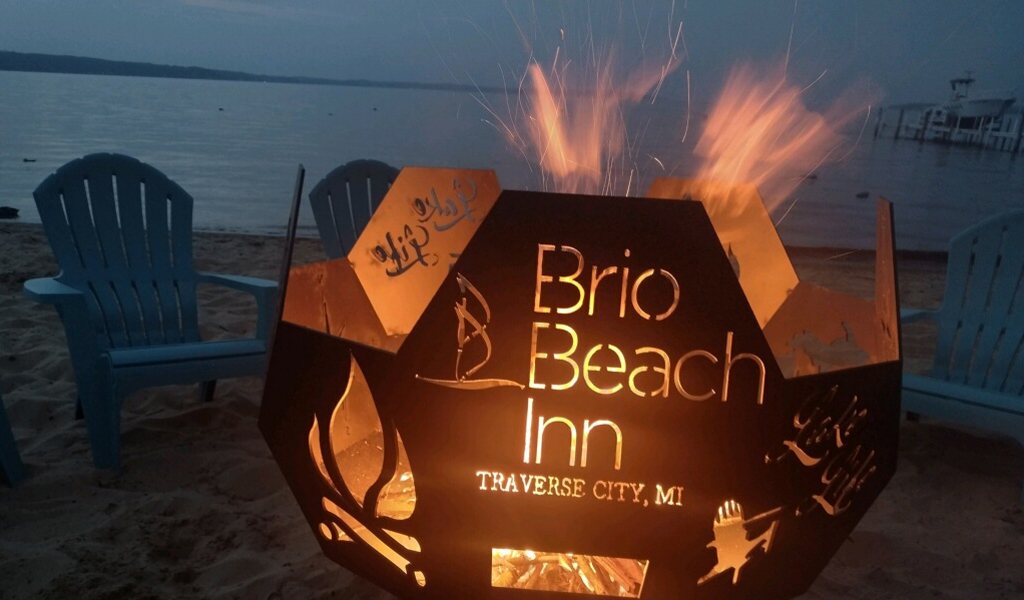 A lit fire pit on a beach with two blue Adirondack chairs and a body of water in the background at dusk.